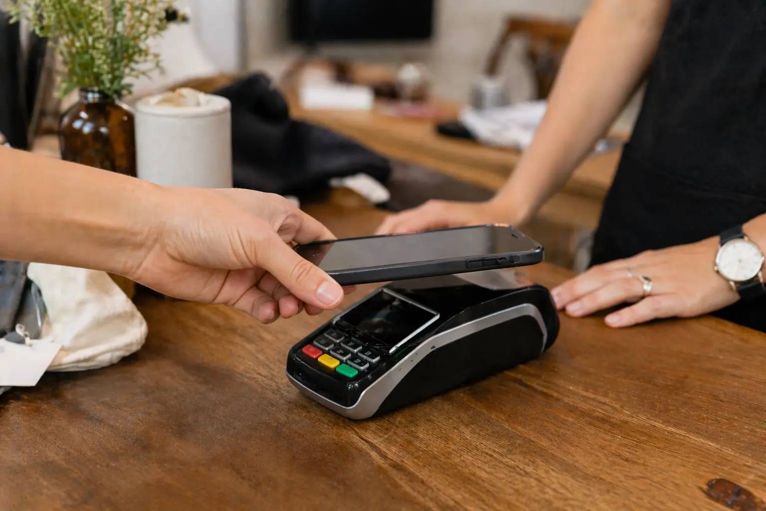 Close-up of a customer tapping a smartphone on a payment terminal at a wooden retail checkout counter in a boutique setting