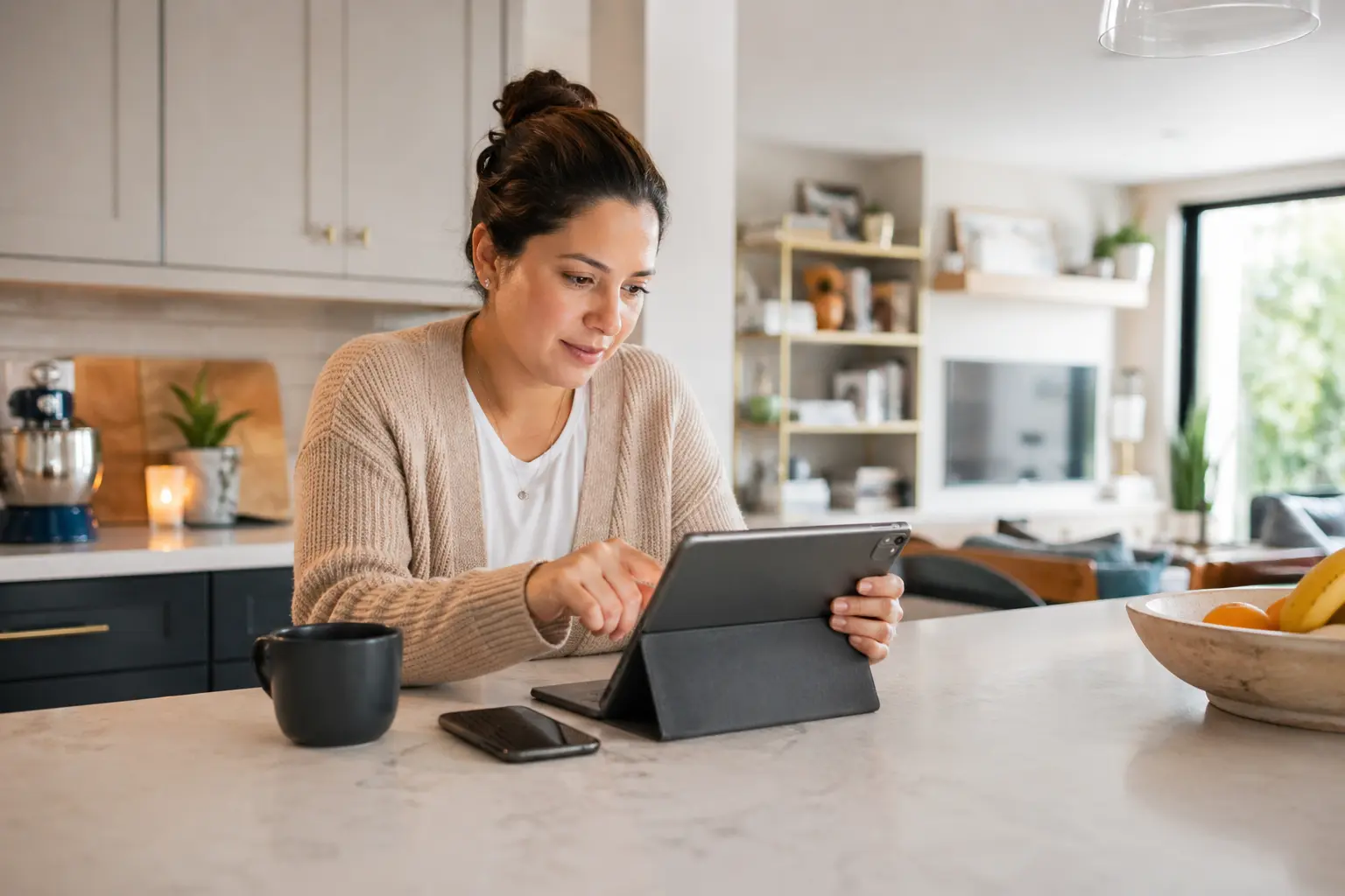 A person sitting at a kitchen island using a tablet and smartphone to manage smart home devices in a modern, lived-in home