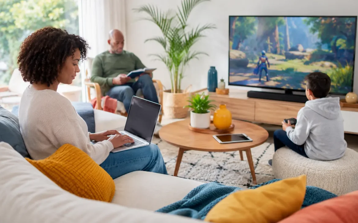 A family uses multiple connected devices in a bright modern living room, including a laptop, video game console, TV, and tablet.