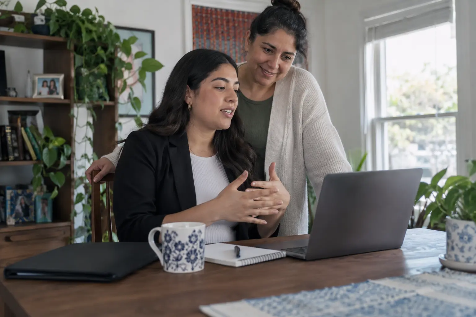 Young adult practices for a virtual job interview on a laptop while a family member offers encouragement.